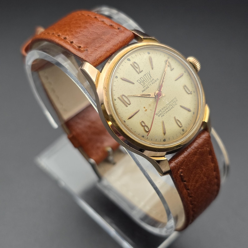 May include: A vintage gold-toned wristwatch with a cream-colored face and brown leather band. The watch face features black numerals and the brand name "Britix". The watch is displayed on a clear stand.