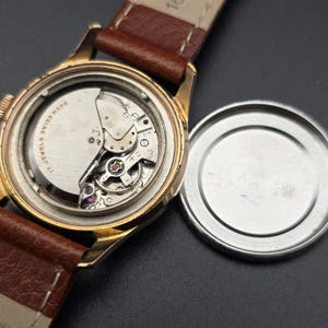 May include: A vintage gold-toned wristwatch with a brown leather strap. The watch's open back reveals the intricate mechanical movement, including gears, jewels, and the inscription "17 JEWELS SWISS MADE". The watch's case is round, and the back cover is detached.