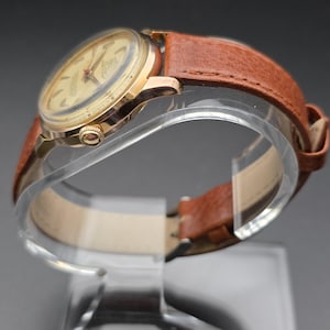May include: A vintage gold-toned wristwatch with a light-colored face and red accents. The watch features a brown leather band. The watch is displayed on a clear stand, showcasing its classic design and craftsmanship.
