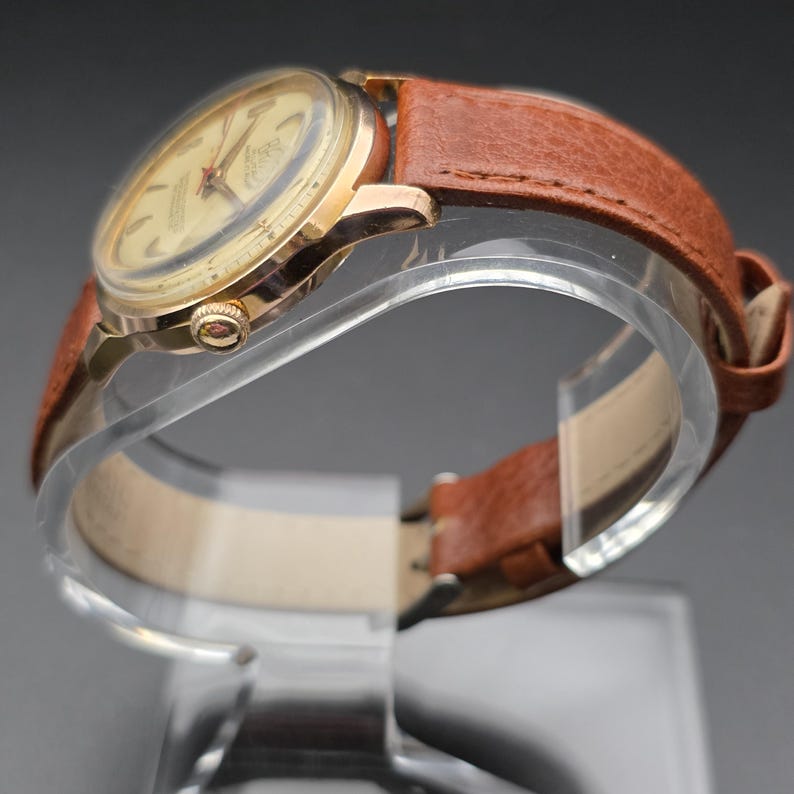 May include: A vintage gold-toned wristwatch with a light-colored face and red accents. The watch features a brown leather band. The watch is displayed on a clear stand, showcasing its classic design and craftsmanship.