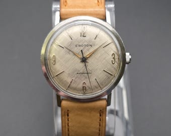 May include: A vintage Croton wristwatch featuring a round silver-tone case and a textured champagne-colored dial with a crosshatch pattern. The dial displays the brand name Croton, the word Waterproof, and gold-tone hands with a tan leather strap.