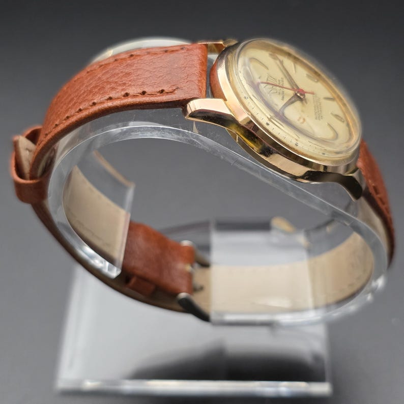 May include: A vintage gold-toned wristwatch with a light-colored face and red accents. The watch features a brown leather band. The watch is displayed on a clear stand, showcasing its classic design and craftsmanship.