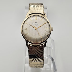 May include: A gold-toned Zodiac Automatic wristwatch with a textured silver face and gold hands. The watch features the text "Zodiac Automatic" and "25 Jewels". The watch has a gold expanding band.