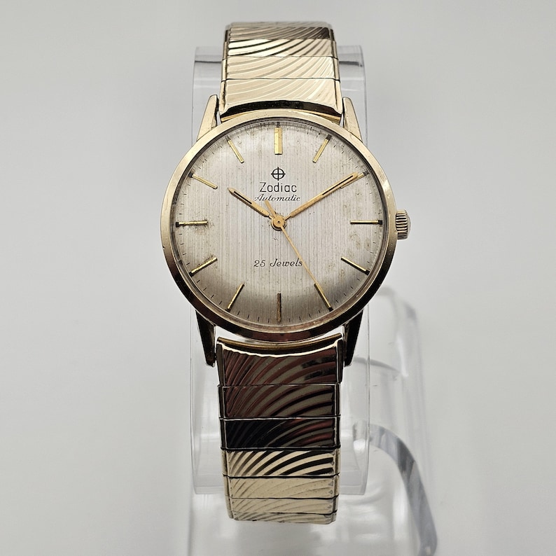 May include: A gold-toned Zodiac Automatic wristwatch with a textured silver face and gold hands. The watch features the text "Zodiac Automatic" and "25 Jewels". The watch has a gold expanding band.