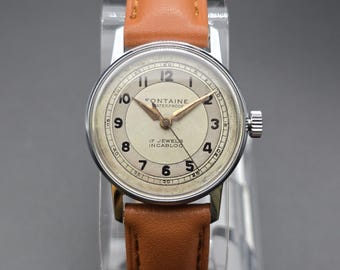 May include: A vintage-style wristwatch with a round silver-tone case and a light tan leather strap. The off-white dial features black Arabic numerals, gold-tone hands, and the text Fontaine Waterproof 17 Jewels Incabloc printed in black.