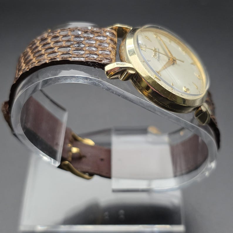 K&ouml;nnte beinhalten: Eine goldfarbene Armbanduhr mit hellem Zifferblatt und goldenen Zeigern. Die Uhr hat ein braunes Lederarmband mit einem strukturierten, gemusterten Abschnitt in der N&auml;he des Zifferblatts. Die Uhr wird auf einem transparenten St&auml;nder pr&auml;sentiert.
