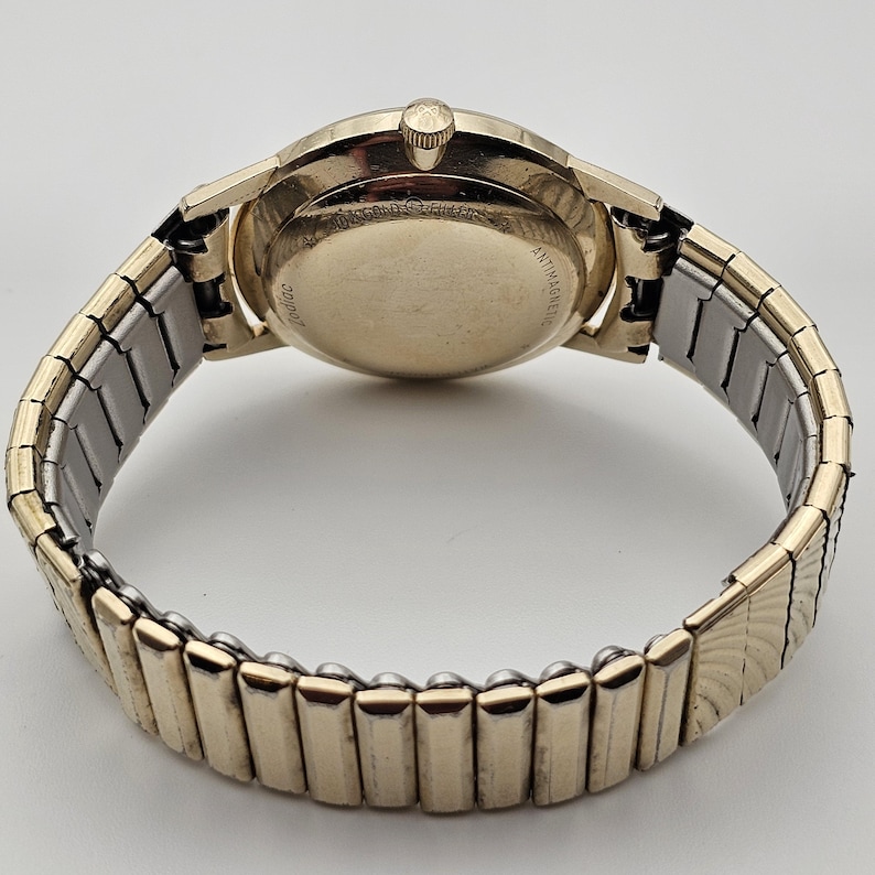 May include: A vintage gold-toned wristwatch with a stretch band. The watch face is round, and the band is a combination of gold and silver-toned metal. The watch is viewed from the back, showing the watch's inner workings and the words "ANTIMAGNETIC" and "ZODIAC".