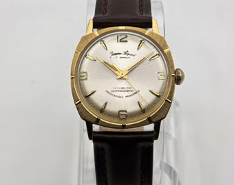May include: A vintage gold-coloured wristwatch with a silver face and brown leather strap. The watch face features gold hands and hour markers, with the text "Supreme Regent" and "Incabloc" visible. The watch is displayed on a clear stand.