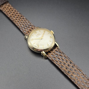 K&ouml;nnte beinhalten: Eine vintage goldfarbene Armbanduhr mit cremefarbenem Zifferblatt und goldenen Zeigern und Ziffern. Die Uhr hat ein braunes, strukturiertes Lederarmband. Das Zifferblatt zeigt den Markennamen. Eine klassische Uhr.