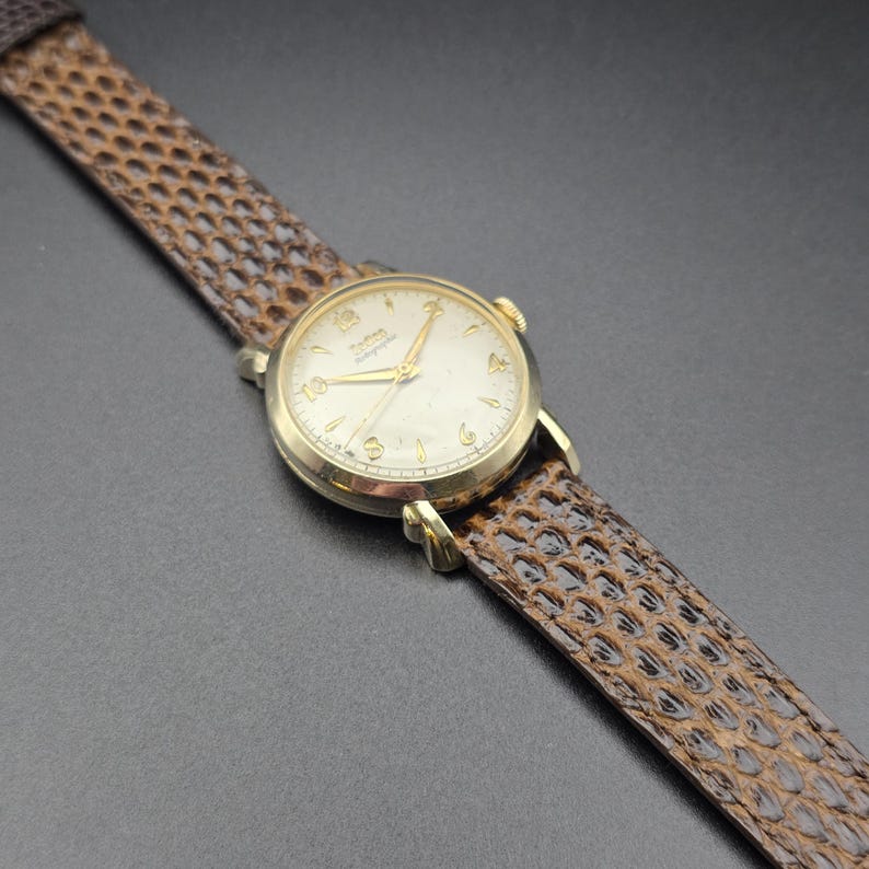 K&ouml;nnte beinhalten: Eine vintage goldfarbene Armbanduhr mit cremefarbenem Zifferblatt und goldenen Zeigern und Ziffern. Die Uhr hat ein braunes, strukturiertes Lederarmband. Das Zifferblatt zeigt den Markennamen. Eine klassische Uhr.