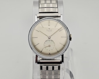 May include: A silver Alpha watch with a white face and black hands. The watch has a silver metal band and says "Alpha 17 Jewels" on the face.
