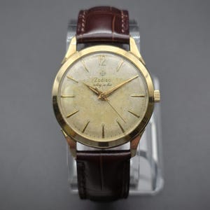 Pode incluir: Um rel&oacute;gio de pulso Zodiac vintage com caixa dourada e bracelete de couro castanho. O mostrador do rel&oacute;gio &eacute; de cor dourada clara com ponteiros e marcadores dourados. O texto "Zodiac" e "rotog-raphic" &eacute; vis&iacute;vel. O rel&oacute;gio tem um design cl&aacute;ssico e elegante.