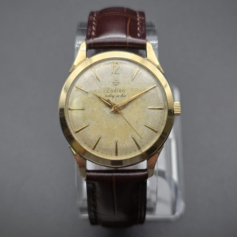 Pode incluir: Um rel&oacute;gio de pulso Zodiac vintage com caixa dourada e bracelete de couro castanho. O mostrador do rel&oacute;gio &eacute; de cor dourada clara com ponteiros e marcadores dourados. O texto "Zodiac" e "rotog-raphic" &eacute; vis&iacute;vel. O rel&oacute;gio tem um design cl&aacute;ssico e elegante.