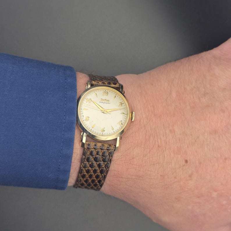 K&ouml;nnte beinhalten: Eine goldfarbene Armbanduhr mit cremefarbenem Zifferblatt und goldenen Zeigern. Die Uhr hat ein braunes, strukturiertes Lederarmband. Die Uhr befindet sich an einem Handgelenk, mit einem blauen &Auml;rmel im Hintergrund.