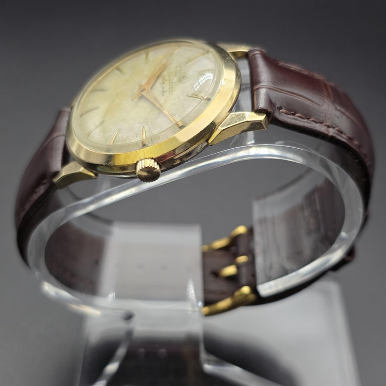Pode incluir: Um rel&oacute;gio de pulso vintage dourado com mostrador claro e ponteiros dourados. O rel&oacute;gio tem uma pulseira de couro castanho com fivela dourada. O rel&oacute;gio &eacute; exibido em um suporte transparente e curvo.