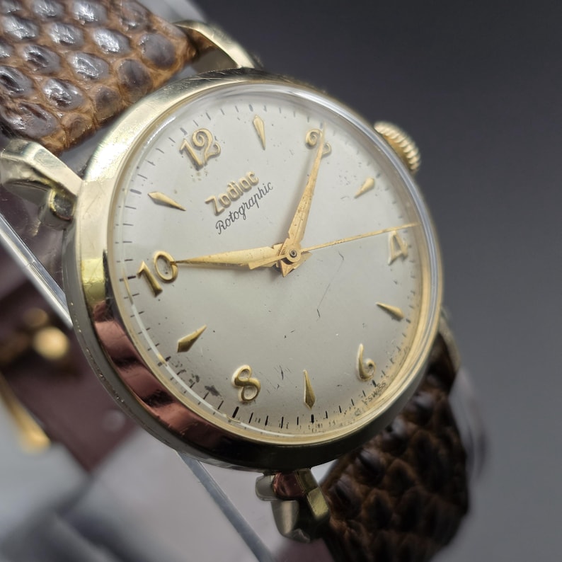 K&ouml;nnte beinhalten: Eine Vintage-Armbanduhr Zodiac Rotographic in Goldoptik mit hellem Zifferblatt und goldenen Zeigern und Markierungen. Die Uhr hat ein braunes Lederarmband mit Reptilienmuster. Das Zifferblatt zeigt die Zahlen 12, 10, 4 und 6.