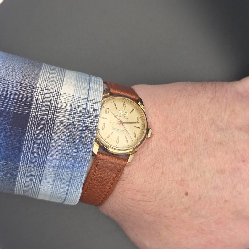 May include: A gold-toned wristwatch with a light-colored face and brown leather band. The watch face displays the brand name and numbers. The watch is worn on a wrist, partially obscured by a blue and white plaid shirt sleeve.