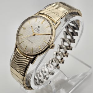 May include: A vintage Zodiac Automatic wristwatch with a gold-toned case and a silver-colored dial. The watch features gold hands and markers, and the text "Zodiac Automatic" and "25 Jewels" are visible on the dial. The watch has a gold-toned, textured band.
