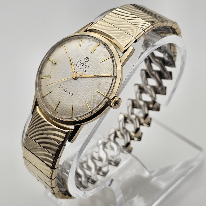 May include: A vintage Zodiac Automatic wristwatch with a gold-toned case and a silver-colored dial. The watch features gold hands and markers, and the text "Zodiac Automatic" and "25 Jewels" are visible on the dial. The watch has a gold-toned, textured band.