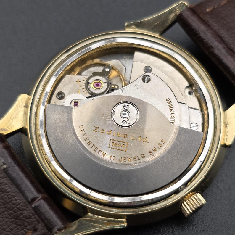 Pode incluir: Vista de perto da parte de tr&aacute;s de um rel&oacute;gio Zodiac Ltd. dourado, revelando o intrincado movimento mec&acirc;nico. O rel&oacute;gio apresenta uma bracelete de couro castanho e o texto "SEVENTEEN 17 JEWELS SWISS". O rel&oacute;gio est&aacute; marcado "1824".