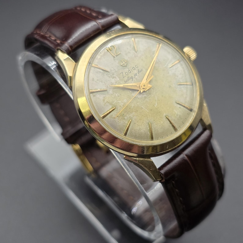 Pode incluir: Um rel&oacute;gio de pulso Zodiac vintage em tom dourado com uma pulseira de couro castanho. O mostrador do rel&oacute;gio &eacute; de cor dourada clara com ponteiros e marcadores dourados. O rel&oacute;gio tem as palavras "Zodiac" e "Seolographic" impressas no mostrador.