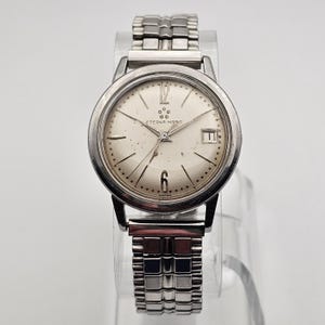 Puede incluir: Un reloj de pulsera Eterna-Matic plateado vintage. El reloj presenta una esfera redonda con una esfera de color claro, manecillas plateadas y una ventana de fecha a las 3 en punto. La correa del reloj está hecha de eslabones de metal plateado. La esfera del reloj muestra el número 12 y el número 6.