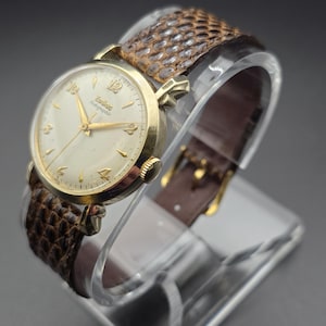 K&ouml;nnte beinhalten: Eine Vintage-Zodiac-Armbanduhr mit goldfarbenem Geh&auml;use und cremefarbenem Zifferblatt. Die Uhr hat goldene Zeiger und Stundenmarkierungen. Sie hat ein braunes, strukturiertes Lederarmband. Die Uhr wird auf einem transparenten St&auml;nder pr&auml;sentiert.