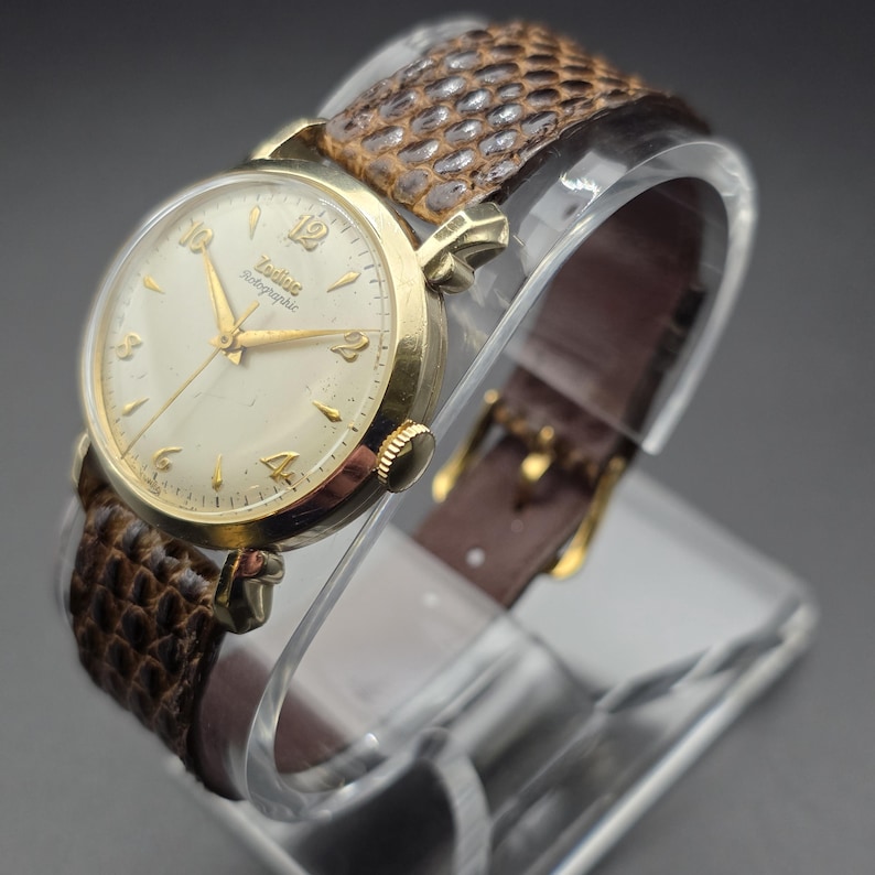 K&ouml;nnte beinhalten: Eine Vintage-Zodiac-Armbanduhr mit goldfarbenem Geh&auml;use und cremefarbenem Zifferblatt. Die Uhr hat goldene Zeiger und Stundenmarkierungen. Sie hat ein braunes, strukturiertes Lederarmband. Die Uhr wird auf einem transparenten St&auml;nder pr&auml;sentiert.