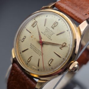 May include: A vintage gold-toned Britix military watch with a light-colored face, black numerals, and a red second hand. The watch has a brown leather strap. The watch face includes the text "Britix Militar Ancre 17 Rubis Superautomatic Shockprotected Antimagnetic".