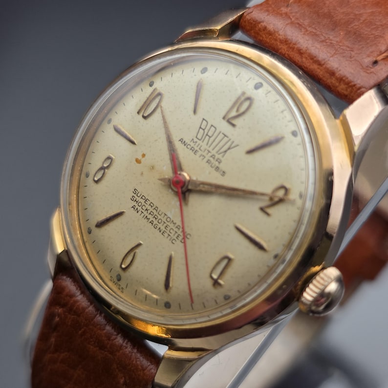 May include: A vintage gold-toned Britix military watch with a light-colored face, black numerals, and a red second hand. The watch has a brown leather strap. The watch face includes the text "Britix Militar Ancre 17 Rubis Superautomatic Shockprotected Antimagnetic".
