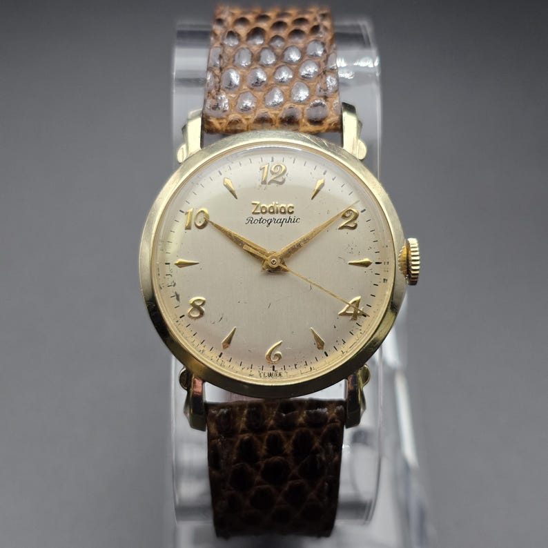 K&ouml;nnte beinhalten: Eine Vintage Zodiac Rotographic Armbanduhr mit goldfarbenem Geh&auml;use und cremefarbenem Zifferblatt. Die Uhr hat goldene Zeiger und Stundenmarkierungen sowie ein braunes Lederarmband mit Schlangenmuster. Die Uhr wird auf einem transparenten St&auml;nder pr&auml;sentiert.