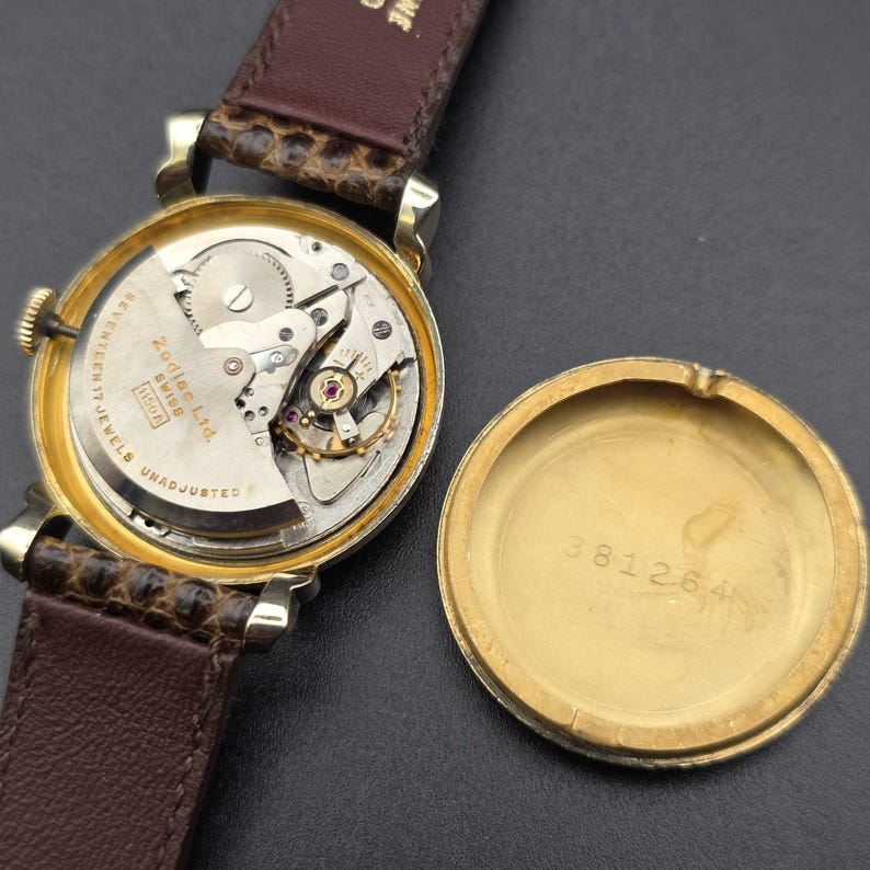 K&ouml;nnte beinhalten: Eine offene Vintage-Zodiac-Uhr mit braunem Lederarmband. Das goldfarbene Geh&auml;use zeigt das Uhrwerk. Das abgenommene Geh&auml;use zeigt die Seriennummer.