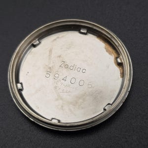 Pode incluir: Parte traseira prateada de um rel&oacute;gio com o nome da marca "Zodiac" e o n&uacute;mero "594006" gravados. N&uacute;meros manuscritos adicionais tamb&eacute;m est&atilde;o presentes. A parte traseira do rel&oacute;gio tem formato circular e apar&ecirc;ncia ligeiramente desgastada.