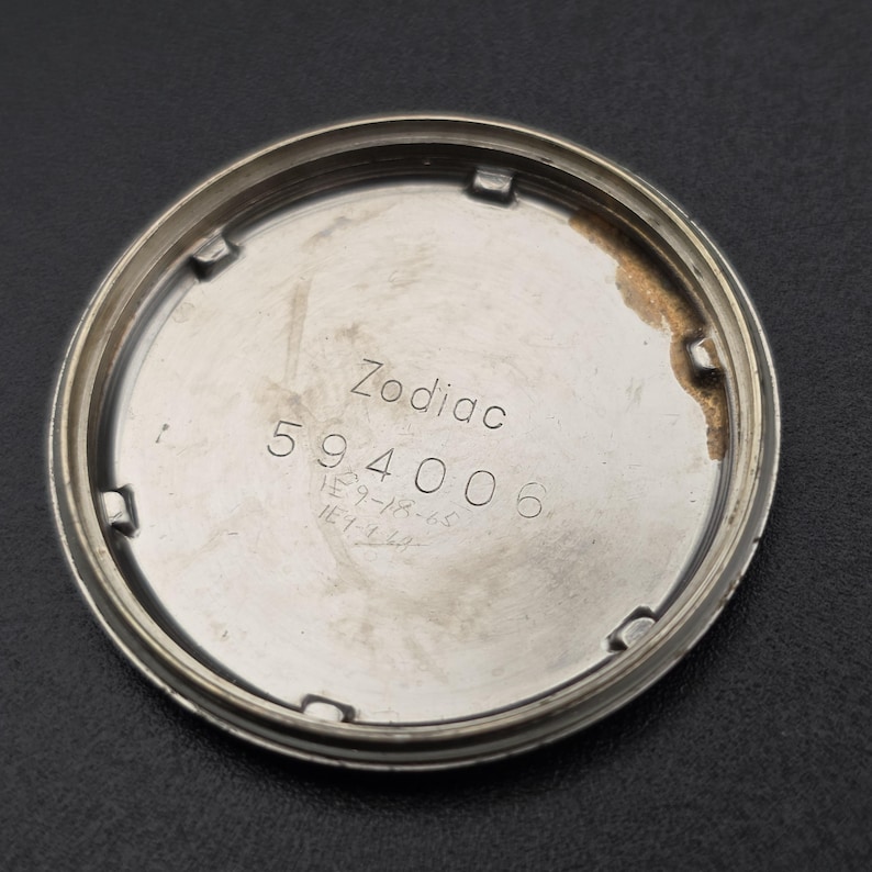 Pode incluir: Parte traseira prateada de um rel&oacute;gio com o nome da marca "Zodiac" e o n&uacute;mero "594006" gravados. N&uacute;meros manuscritos adicionais tamb&eacute;m est&atilde;o presentes. A parte traseira do rel&oacute;gio tem formato circular e apar&ecirc;ncia ligeiramente desgastada.
