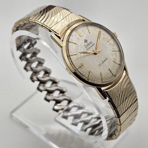 May include: A gold-toned Zodiac Automatic watch with a textured silver face and gold hands. The watch features the text "Zodiac Automatic" and "25 Jewels". The watch band is gold-toned with a textured design.