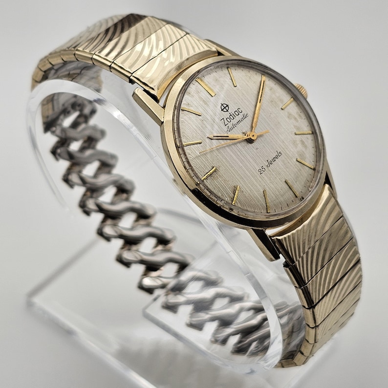 May include: A gold-toned Zodiac Automatic watch with a textured silver face and gold hands. The watch features the text "Zodiac Automatic" and "25 Jewels". The watch band is gold-toned with a textured design.