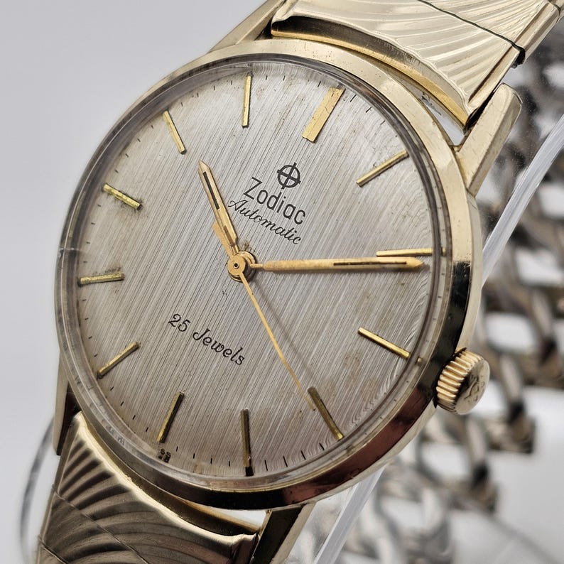 May include: A vintage Zodiac Automatic wristwatch with a gold-toned case and band. The watch face is silver with gold hands and markers. The text "Zodiac Automatic" and "25 Jewels" are visible on the watch face. The watch has a textured, brushed metal finish.
