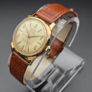 May include: A vintage gold-toned wristwatch with a cream-colored face and brown leather band. The watch face features black numerals and the brand name "Britix". The watch is displayed on a clear stand.