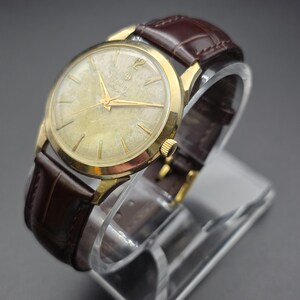 Pode incluir: Um rel&oacute;gio Zodiac vintage com caixa dourada e pulseira de couro castanho. O mostrador do rel&oacute;gio &eacute; dourado claro com ponteiros e marcadores dourados. A pulseira &eacute; castanho escuro com um padr&atilde;o texturizado. O rel&oacute;gio &eacute; exibido em um suporte transparente.
