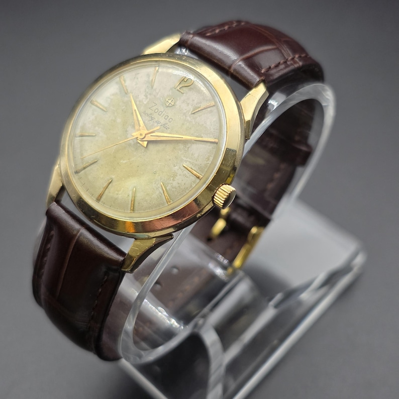 Pode incluir: Um rel&oacute;gio Zodiac vintage com caixa dourada e pulseira de couro castanho. O mostrador do rel&oacute;gio &eacute; dourado claro com ponteiros e marcadores dourados. A pulseira &eacute; castanho escuro com um padr&atilde;o texturizado. O rel&oacute;gio &eacute; exibido em um suporte transparente.