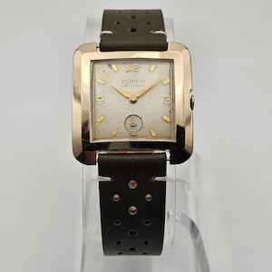 Op de afbeelding: Een vintage Benrus polshorloge met een goudkleurige vierkante kast en een gespikkelde, lichtgekleurde wijzerplaat. Het horloge heeft gouden wijzers en markeringen, met het nummer 12 bovenaan. De donkerbruine leren band heeft een geperforeerd ontwerp.