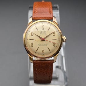 May include: A vintage gold-toned wristwatch with a brown leather band. The watch face is cream-colored with black numerals and hands, and a red second hand. The watch has the text "BRITIX" and "SUPERAUTOMATIC SHOCKPROTECTED ANTIMAGNETIC" on the face.