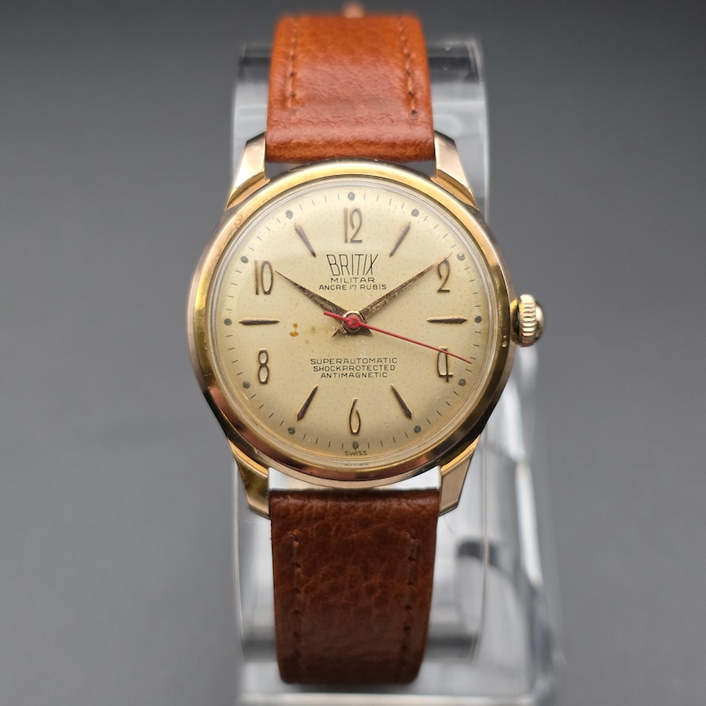 May include: A vintage gold-toned wristwatch with a brown leather band. The watch face is cream-colored with black numerals and hands, and a red second hand. The watch has the text "BRITIX" and "SUPERAUTOMATIC SHOCKPROTECTED ANTIMAGNETIC" on the face.