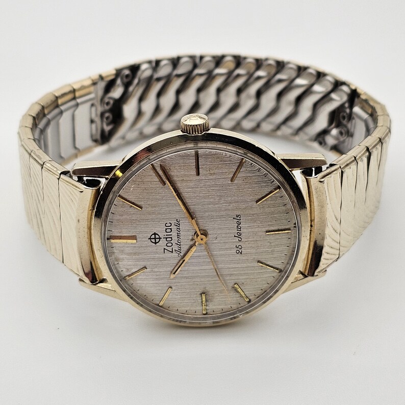 May include: A vintage Zodiac Automatic wristwatch with a silver-tone dial and gold-tone hands and markers. The watch features a gold-tone bezel and a stainless steel expansion band. The dial reads "Zodiac Automatic" and "25 Jewels".