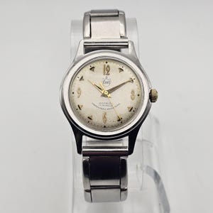 May include: A silver wristwatch with a white face and black hands. The watch has a gold-colored logo that says "Cort" and the text "Incabloc 17 Jewels Unbreakable Mainspring".