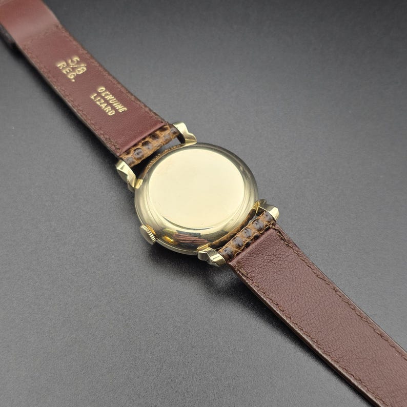 K&ouml;nnte beinhalten: Nahaufnahme einer goldfarbenen Uhr mit braunem Lederarmband. Das Zifferblatt der Uhr ist kreisf&ouml;rmig und poliert. Das Armband ist aus echtem Echsenleder und 1,6 cm breit.