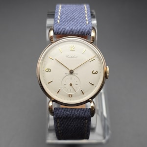 May include: A vintage gold-toned wristwatch with a cream-colored face, featuring gold hands and numerals. The watch has a small seconds dial and is marked "Contbert" and "Antimagnetic." It is attached to a blue denim strap with gold stitching.