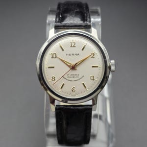 Puede incluir: Un reloj de pulsera HERNA plateado vintage con esfera crema, manecillas doradas y correa de cuero negro. La esfera del reloj muestra el texto "HERNA", "17 JEWELS" e "INCABLOC". El reloj tiene un diseño clásico.