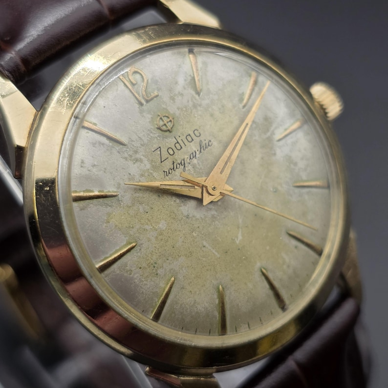 Pode incluir: Rel&oacute;gio Zodiac rotographic vintage com caixa dourada e bracelete em couro castanho. O mostrador do rel&oacute;gio &eacute; dourado desbotado com ponteiros e marcadores dourados. O texto "Zodiac rotographic" est&aacute; impresso no mostrador.