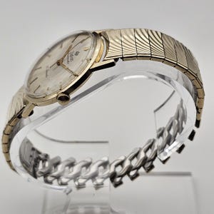 May include: A vintage Zodiac automatic wristwatch with a gold-toned case and a silver-colored dial. The watch features gold hands and hour markers, and a gold-toned expanding band. The watch is displayed on a clear stand.